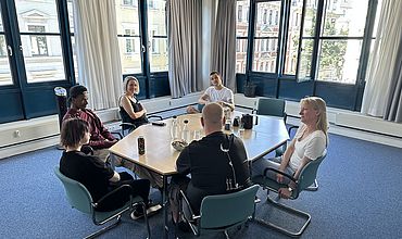 Mitarbeitende der Jugendberufshilfe sitzen gemeinsam mit Heike Praetz, stv. Geschäftsführerin des Jobcenters Leipzig, an einem Tisch. Alle Personen scheinen in ein anregendes Gespräch vertieft zu sein. Der Teppich ist blau, die Fenster im Hintergrund sind teilweise geöffnet.