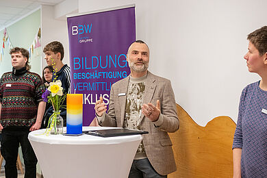 Mittig im Bild Dr. Ronny Valdorf an einem Stehtisch. Auf dem Tisch steht eine bund gefärbte Kerze und ein kleiner Blumenstrauß. im hintergrund das Roll-Up der BBW-Leipzig-Gruppe. Valdorf scheint gerade eine Rede zu halten. 3 Personen stehen am Rand des Bildes und hören zu.
