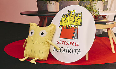 Links Eulen-Kuscheltier und daneben ein großes rundes Holzschild mit Logo und Aufschrift "Gütesiegel Buchkita"