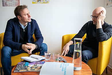 Zwei Personen sitzen sich in gelben Sesseln gegenüber und sprechen miteinander. Auf dem Tisch liegen Broschüren, ein Notizbuch und Gläser mit Wasser.