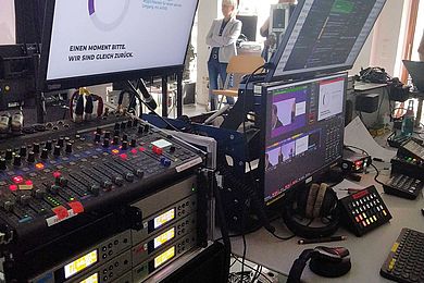 Studioszene aus dem Hintergrund. Es sind zahlreiche Monitore und Regler zu sehen. An den Monitoren vorbeischauend sieht man vor dem Fenster eine Dozentin stehen. 