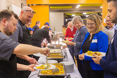 Heißstrecke mit vegetarisch-veganen Hauptgerichten. Auf der linken Seite Mitarbeiter der Diakonischen Unternehmensdienste, in der Mitte die Gerichte, rechts lächelnde Gäste