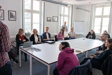 Um einen großen Tisch herum sitzen viele Menschen und unterhalten sich und lachen.