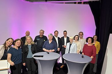 Studioszene mit 13 mitwirkenden Personen des AVWS-Fachtages. Alle lächeln in die Kamera. 