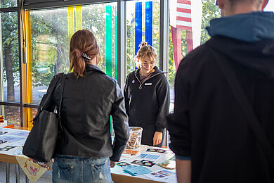 Zwei Personen stehen an einem Informationstisch mit verschiedenen Flyern und Informationsblättern. Hinter dem Tisch im Foyer des Berufsbildungswerks Leipzig steht eine*r Mitarbeiter*in und blickt auf das Informationsmaterial.