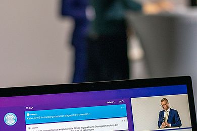 Prof. Dr. Michael Fuchs ist auf einem Bildschirm zu sehen. Eine Gebärdensprachdolmetscherin übersetzt das Gesagte.