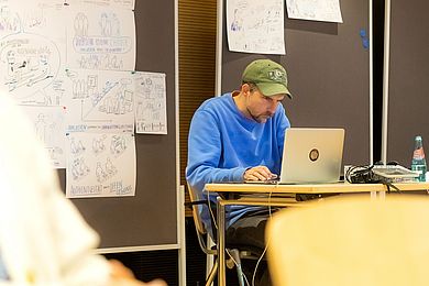 Der Illustrator sitzt an einem Tisch vor einem Laptop, hinter ihm hängen Illustrationen an Pinnwänden
