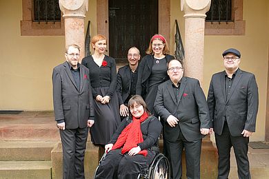 Mitglieder des inklusiven A-Capella-Ensembles Thonkunst vor der St. Gangolf-Kirche in Kohren-Salis. Alle tragen schwarze Kleidung mit roten Accessoires. 