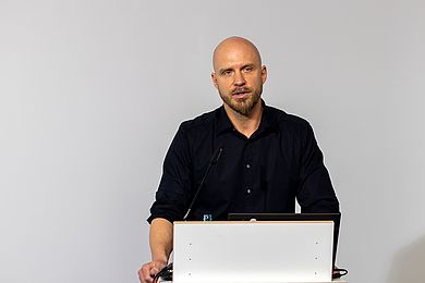 Dr. Oliver Zetsche ist Mitarbeiter der Abteilung Forschung und Entwicklung der BBW-Leipzig-Gruppe. Er spricht am Pult und trägt eine Glatze, einen Vollbart und ein schwarzes Hemd.