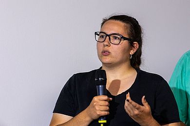 Auf dem Bild sieht man Annabell, die eine Ausbildung als Gärtnerin beim des Berufsbildungswerk Leipzig macht. Sie spricht in ein Mikrofon. Ihre Fingernägel sind schwarz lackiert. Sie trägt ein schwarzes T-Shirt und eine schwarz umrahmte Brille.