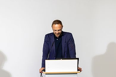 Tobias Schmidt, Hauptgeschäftsführer BBW-Leipzig-Gruppe, steht an einem Pult und spricht in ein schmales, schwarzes Mikrofon. Unter seinem blauen Sakko trägt er ein schwarzes Polohemd. Während seiner Rede schaut er über die hell gerahmte Brille hinweg.