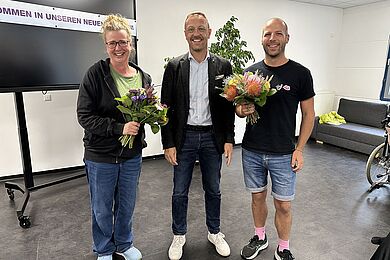 Drei Personen nebeneinander. Zwei halten bunte Blumensträuße. Alle Personen lachen in die Kamera.