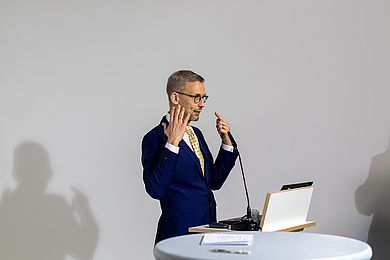Zu sehen ist eine Seitenaufnahme von Prof. Dr. Michael Fuchs, wie er am Pult beim Sprechen mit den Händen gestikuliert.