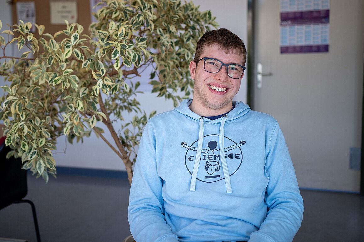 Teilnehmer*in in einem hellblauen Pullover in einem Innenraum. Der Teilnehmer trägt eine Brille und hat kurze braune Haare. Er lächelt breit in die Kamera.