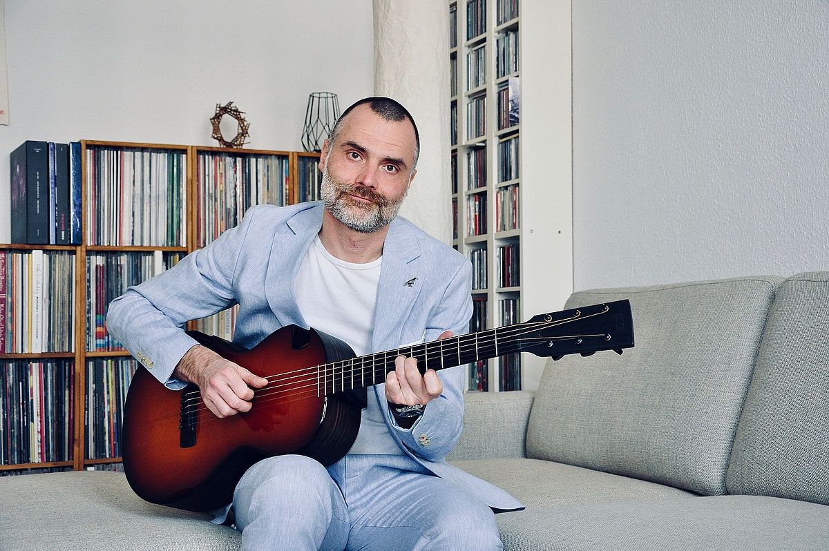 Der Musiker Ronny Valdorf sitzt auf einem Sofa und spielt eine Akusitik-Gitarre. Er trägt einen hellblauen Anzug.