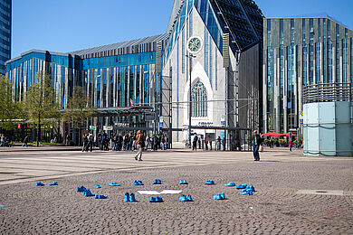 Mehrere Paare leuchtend blauer Schuhe stehen auf dem gepflasterten Boden. Im Hintergrund ist das Augusteum der Universität Leipzig zu sehen.