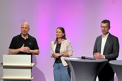 Studioszene mit 2 Dozenten und der Moderatorin in der Mitte am Stehtisch. Alle blicken in die Kamera. 