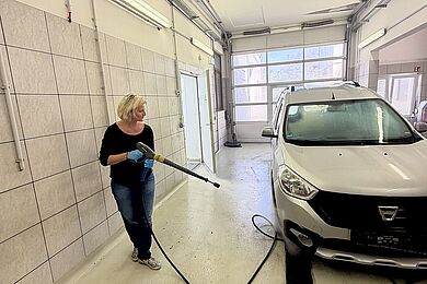 Eine Frau ist in einer Werkstatt und hält einen Hochdruckreiniger aus dem gerade Wasser spritzt. Damit reinigt sie die Felgen eines Wagens.