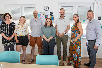 In einem Raum stehen sieben Personen nebeneinander und posieren für ein Gruppenbild, darunter Patrick Schreiber, Präsident des Landesamtes für Schule und Bildung, und Marion Nagel vom Amt für Schule.
