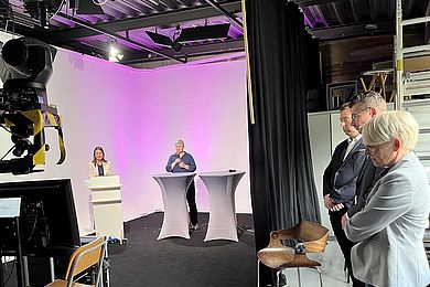 Zweigeteilte Studioszene des AVWS-Fachtages, zwei Referierende stehen links und sprechen in die Kamera. Rechts stehen 3 weitere Referierende und schauen auf einen Monitor. . 