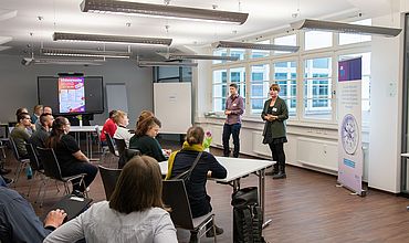 In einem Raum stehen zwei Personen vor einem Publikum, das Publikum sitzt.