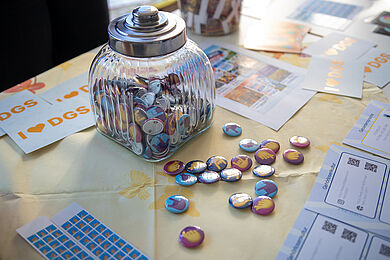 Auf einem Tisch mit Informationsmaterial steht ein großes Glas mit verschiedenen, bunten Buttons. Die Buttons sind auch vor dem Glas auf dem Tisch verteilt. Auf den Buttons sind Hände mit verschiedenen Gebärden abgebildet.