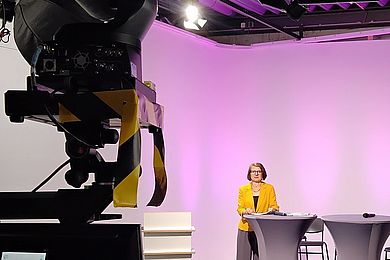 Studioszene des AVWS_Fachtages aus dem Hintergrund fotografiert. Die Moderatorin steht im Hintergrund an Stehtischen und schaut in die Kamera. Links ist die Kamera von hinten zu sehen.