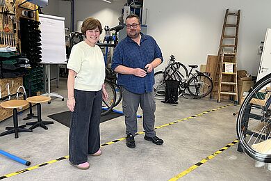 Ein Mann mit einer Brille und eine Frau stehen nebeneinander in einer Fahrradwerkstatt lächeln freundlich in die Kamera. 