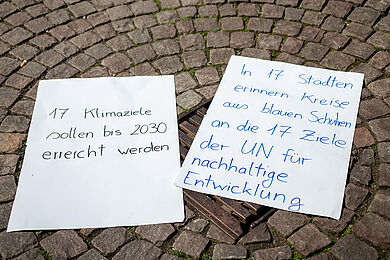 Zwei handbeschriebene Plakate liegen auf gepflastertem Boden. Auf einem Plakat steht "17 Klimaziele sollen bis 2030 erreicht werden", auf dem anderen "In 17 Städten erinnern Kreise aus blauen Schuhen an die 17 Ziele der UN für nachhaltige Entwicklung".