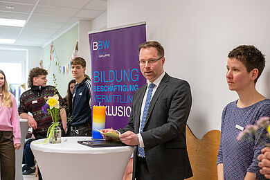 Bastian Schmiedel in einem dunklen Anzug, hält an einem Stehtisch stehend eine Rede. Rechts neben ihm Lea Beuter, die zuhört. Links das Roll-Up der BBW-Leipzig-Gruppe. Links davon mehrere junge Menschen, die ebenso zuhören