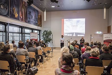 In einem Raum sitzen viele Menschen, die von hinten zu sehen sind. An einer Leinwand wird ein Bild projiziert, vor dieser steht eine Person, die spricht