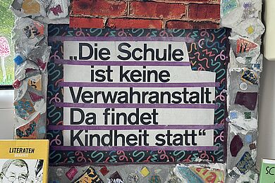 Gerahmtes Kunstobjekt auf einer Fensterbank mit der Aufschrift: „Die Schule ist keine Verwahranstalt. Da findet Kindheit statt.“ Der Rahmen ist mit bunten Mosaikelementen gestaltet.