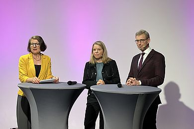 Die Moderatorin rechts im Bild schaut gemeinsam mit den beiden Dozentinnen des Spotlights 2 auf den Bildschirm mit den Zuschauerfragen. Alle Personen stehen an Stehtischen.