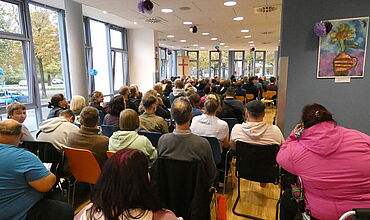 Werkstattmitarbeitende sitzen bei der Jubiläumsfeier auf Stühlen und lauschen den Redebeiträgen.