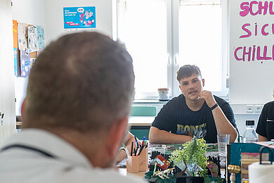 links vorne ist ein Kopf unscharf von hinten zu erkennen. Dem gegenüber sitzt mit Blick Richtung Kamera ein junger Mann, der gerade zu sprechen scheint. Dahinter ein Fenster.