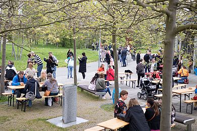 Vor dem Freizeithaus des Berufsbildungswerkes stehen viele Biertischgarnituren, einige Menschen sitzen dort und stehen und unterhalten sich.