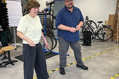 Ein Mann mit einer Brille und eine Frau stehen nebeneinander in einer Fahrradwerkstatt und lächeln. 