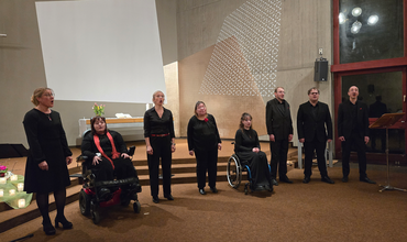 Das Vocalensemble Thonkonst beim Konzert in der Kirche in Esslingen. Die Mitglieder stehen/sitzen im Rollstuhl und singen. Im Hintergrund ist ein sehr moderner Kirchenraum zu sehen mit Elementen aus Holz und Glas.