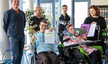 Zwei Personen sitzen im Rollstuhl, hinter ihnen stehen vier weitere Personen. Die linke Person im Rollstuhl lacht in die Kamera. Sie trägt einen hellblauen Hoodie und eine braune Hose. Sie hält eine Rose in der einen Hand und ein Zertifikat der IHK in der anderen. Die rechte Person im Rollstuhl blickt ebenfalls in die Kamera. Sie trägt ein bunt geblümtes Kleid über einem weißen Langarmshirt und einer dunklen Strumpfhose. Die stehende Person neben ihr hält ein weiteres Zertifikat und eine Rose in die Kamera und blickt zur Person im Rollstuhl. Alle Personen auf dem Bild lachen oder blicken in die Kamera, die Stimmung ist fröhlich.