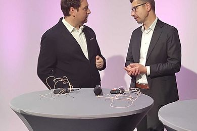 2 Dozenten der Expertinnenrunde unterhalten sich an einem Stehtisch im studio des AVWS-fachtages. Auf dem Stehtisch liegen Mikrofone. 