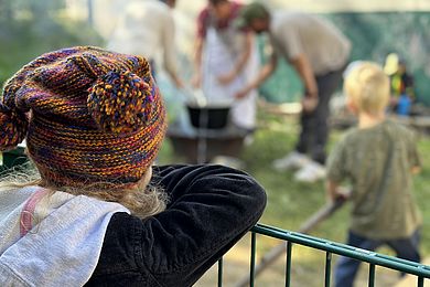 Ein Kind steht mit dem Rücken zur Kamera gekehrt im Vordergrund des Bildes. Es trägt eine Wollmütze mit zwei Bommeln. Im Hintergrund sieht man mehrere Personen vor einem Kochtopf stehen, der über einer Feuerschale erwärmt wird.