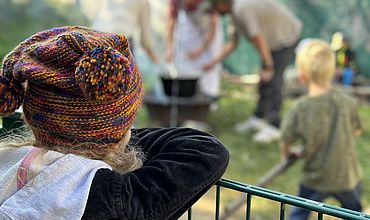 Ein Kind steht mit dem Rücken zur Kamera gekehrt im Vordergrund des Bildes. Es trägt eine Wollmütze mit zwei Bommeln. Im Hintergrund sieht man mehrere Personen vor einem Kochtopf stehen, der über einer Feuerschale erwärmt wird.