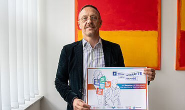Tobias Schmidt mit einem Plakat der Aktionswoche des BeB. Er trägt ein dunkles Jackett über einem gestreiften Hemd. Hinter ihm sind links zugezogene vor einem Fenster zu erkennen und daneben ein Bild mit gelben und roten Colorblocks.