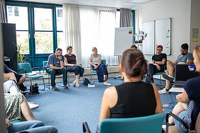 In einem Kreis sitzen mehrere Personen auf Stühlen in einem Büroraum. Unter ihnen Sozialbürgermeisterin Dr. Martina Münch. Im Hintergrund sind Whiteboards zu sehen.