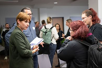 Im Vordergrund sieht man eine Gruppe von vier Personen, die sich unterhält, im Hintergrund sieht man verschwommen weitere Personen.