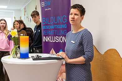 Lea Beuter steht rechts im Bild an einem Stehtisch und hält eine Ansprache. Im Hintergrund ist das Roll-Up der BBW-Leipzig-Gruppe zu erkennen. Daneben stehen junge Leute und hören zu.