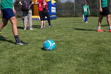 Ein blauer Ball liegt auf einer Wiese. Ein Fuß setzt zum Stoß an. Im Hintergrund sind einige Zuschauer*innen zu sehen.