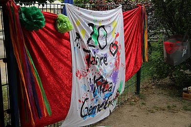 An einem Zaun hängt ein Transparent mit der Aufschrift: 20 Jahre Kita Gerne Groß. Diese ist umrahmt von bunten Farbklecksen.
