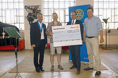 Vier Männer stehen nebeneinander in einem hellen Raum mit großen Fenstern. Zwei von ihnen halten gemeinsam einen übergroßen Spendenscheck des Rotary Clubs in der Hand. Auf dem Scheck ist ein Betrag von 6.000 Euro zu sehen, bestimmt für ein Projekt namens „Diakonie am Thonberg – Inklusive Klassenfahrt“. Im Hintergrund sind ein Banner des Rotary Clubs, moderne Kunstwerke sowie Pflanzen und Sitzmöbel zu erkennen. Die Stimmung wirkt freundlich und formell.