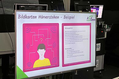 Es ist ein Präsentationsmonitor im Studio des 8. AVWS-Fachtages sichtbar, auf dem der Inhalt "Bildkarten Hörverstehen" mit 2 pink illustrierten quadratischen Bildern dargestellt ist. Dieses Bild stammt aus dem Spotlight 4. 